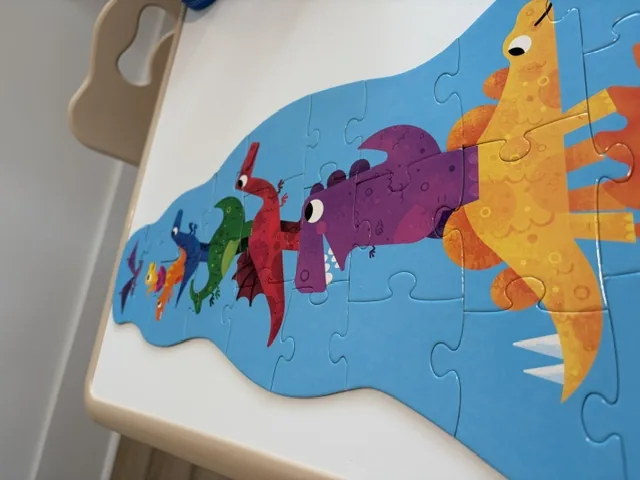 A nearly finished children's dinosaur puzzle on a white play table, with an orange stegosaurus, a purple dinosaur, a red dinosaur, and a green dinosaur stacked on top of each other